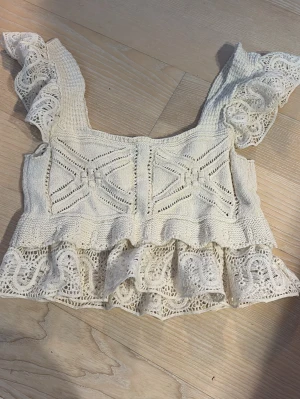Vit stickad topp med spetsvolang - Superfin vit stickad topp med breda axelband och dekorativa spetsvolanger både vid axlarna och nederkanten. Snygga stickade detaljer och mönster framtill ger en boho-vibe. Perfekt för sommardagar och festivaler!
