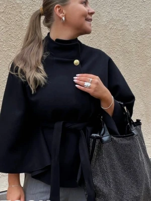 Svart missäy poncho - Snygg svart kappa med breda ärmar och högt krage, knyts i midjan med bälte för en cool siluett. Guldknapp vid halsen ger extra stilpoäng. Perfekt för dig som gillar minimalistisk och chic look.