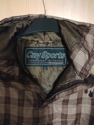 Brun rutig vindjacka från Cny Sports - Säljer en brun och beige rutig vindjacka från Cny Sports i storlek L. Jackan har huva med snörning, dragkedja framtill och är tillverkad i ett lätt syntetmaterial. Perfekt för dig som gillar streetwear och vill ha en unik look.