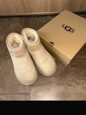 Beige UGG boots med fårskinn - Säljer ett par klassiska UGG boots i beige färg med mjukt fårskinnsfoder och rund tå. Skorna har platt sula med UGG-logga undertill och är tillverkade i mocka. Perfekta för kalla dagar och har en enkel, tidlös design.