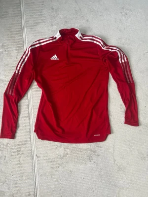 Röd Adidas långärmad träningströja - Säljer en röd långärmad träningströja från Adidas med vita ränder längs axlar och ärmar, samt vit Adidas-logga på bröstet. Tröjan har en halv dragkedja framtill. Perfekt för sport och aktivitet.