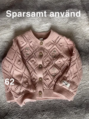 Rosa stickad kofta från Newbie - Superfin rosa stickad kofta från Newbie med dekorativa hålmönster och broderade blommor. Koftan har rund hals, ribbade muddar och träknappar framtill. Perfekt för mysiga dagar och passar till många outfits.🌸 Sparsamt använd. Storlek 62