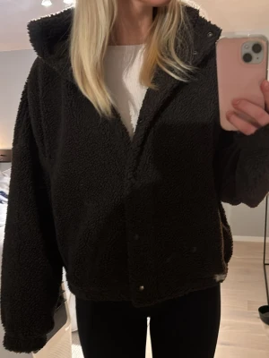 Brun teddyjacka med hög krage - Mysig mörk brun teddyjacka med hög krage och tryckknappar framtill. Jackan har en oversized passform och är tillverkad i fluffigt teddy-material som håller dig varm. Perfekt för kyliga dagar och enkel att styla med dina favoritjeans eller leggings.