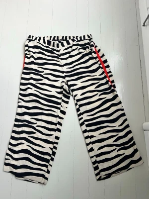 Zebramönstrade wide pants med röd detalj - Säljer ett par sjukt snygga wide pants med zebramönster i svart och vitt. Byxorna har en röd rand längs sidan och elastisk midja för skön passform. Materialet känns mjukt och lätt, perfekt för dig som vill sticka ut med din stil.