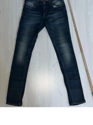 Mörkblå skinny jeans från Nudie - Säljer ett par mörkblå skinny jeans från Nudie med snygga slitningar och tvättade detaljer. Klassisk femficksmodell med Nudie-logga på bakfickan och läderpatch i midjan. Jeansen har smal passform och är tillverkade i stretchig bomull för extra komfort.