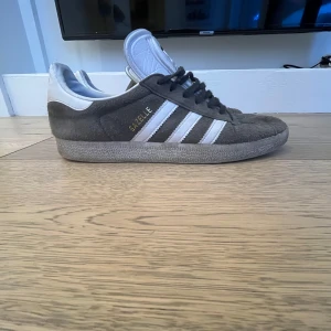 Adidas Gazelle grå sneakers - Säljer ett par klassiska Adidas Gazelle sneakers i grå mocka med vita detaljer och svarta snören. Skorna är synligt använda, men inga hål eller skador. 