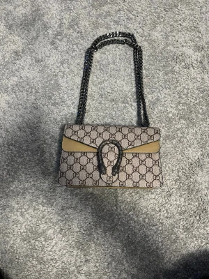 Beige GG axelväska med kedja - Säljer en beige axelväska.Väskan har en snygg kedjerem i metall och ett dekorativt spänne framtill. Perfekt storlek för det viktigaste och ger en lyxig touch till din outfit.