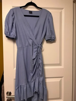 Ljusblå omlottklänning med volang - Supersöt ljusblå omlottklänning från Gina Tricot med puffiga kortärmar, knyt i midjan och volangkant nertill. Klänningen har v-ringning och är perfekt för vår och sommar. 