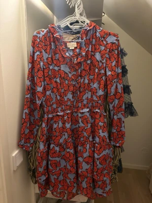Mönstrad kortklänning från Holly & Whyte - Säljer en färgstark kortklänning från Holly & Whyte i storlek S. Klänningen har ett blått tyg med ett iögonfallande rött fjärilsmönster, volangdetaljer och smock i midjan. Långärmad och med v-ringad hals. Perfekt för dig som gillar unika prints och bekväm passform. Den är verkligen så himla vacker och unik älskar verkligen denna kläning.