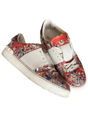Valentino Garavani blommiga sneakers - Säljer ett par Valentino Garavani sneakers med blommigt mönster i rött, vitt, blått och gult. Skorna har vita snören, platt sula och coola nitar vid hälen. Materialet är skinn och de har en rund tå. Perfekt för dig som gillar statement pieces och designer.