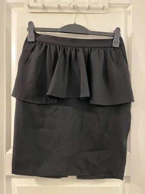 Svart volangkjol från Monki - Säljer en svart kjol från Monki i storlek M. Kjolen har en kort passform och är dekorerad med en bred volang upptill som ger en cool och trendig look. Materialet känns mjukt och har en lätt struktur, perfekt för att styla med sneakers eller boots.