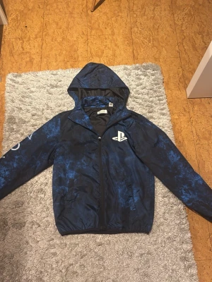Blå PlayStation vindjacka  - Säljer en blå och svart PlayStation vindjacka från H&M med coolt mönster och stora PlayStation-loggor på bröstet, ryggen och ena ärmen. Jackan har huva, dragkedja och två fickor framtill. Perfekt för dig som älskar gaming och vill sticka ut.