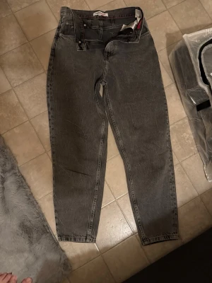 Grå jeans från Tommy Jeans - Säljer ett par grå jeans från Tommy Jeans med klassisk femficksdesign och snygga röda detaljer på insidan. Jeansen har raka ben, normal passform och tydlig logga bak på linningen. Materialet är robust denim i bomull.