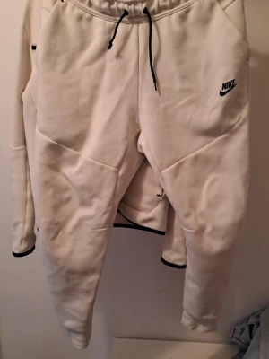 Vita mjukisbyxor från Nike, storlek M - Säljer ett par vita mjukisbyxor från Nike i storlek M. Byxorna har svarta detaljer som snörning i midjan och logga på låret. Tillverkade i mjukt material med ribbade muddar vid benslut och dragkedja på ena bakfickan. Perfekta för chill eller träning.