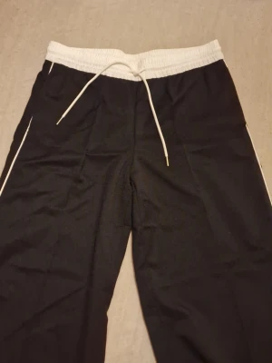 Svarta mjukisbyxor från H&M Divided S - Svarta mjukisbyxor från H&M Divided i storlek S. Byxorna har vita detaljer längs sidorna, vit resårmidja och snörning i midjan. Sköna och loose fit med raka ben, perfekta för chill dagar eller träning. Gillar du baggy kläder? Då är detta perfekt för dig som gillar att vara modest under ditt träningspass. Priset som står gäller, kan möjligtvis diskuteras 🫶