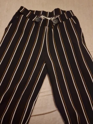 Randiga byxor från Isolde, storlek L - Säljer ett par svarta byxor från Isolde i storlek L med vita och bruna vertikala ränder. Byxorna har hög midja, stora svarta knappar framtill och ett mjukt, stretchigt material. Perfekta för dig som gillar en trendig och bekväm look. Priset som står gäller, kan möjligtvis diskuterats🫶