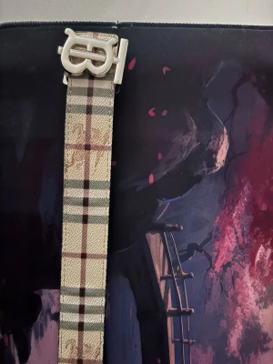 Rutig Burberry bälte med metallspänne - Säljer ett snyggt Burberry bälte med klassiskt rutig design i beige, brunt och svart. Bältet har ett stort metallspänne med Burberrys ikoniska B-logga. Materialet är skinn med präglad yta och insidan är svart. Perfekt för att lyfta din outfit.