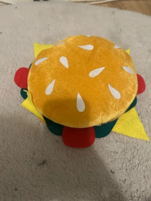 Rolig hamburger hatt i plysch - Unik hatt formad som en hamburgare med sesamfrödetaljer på ovansidan. Hatten har färgglada inslag i gult, rött, grönt och vitt som liknar ost, sallad och tomat. Tillverkad i mjuk plysch och filtmaterial, perfekt för dig som vill sticka ut.