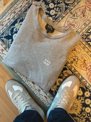 Grå sweatshirt från A.P.C. - Säljer en stilren grå sweatshirt från A.P.C. med vit text 'A.P.C. 19-87' tryckt på bröstet. Tröjan har rund halsringning och långa ärmar, perfekt för en clean och avslappnad look. Materialet är mjukt och skönt, troligtvis bomull.
