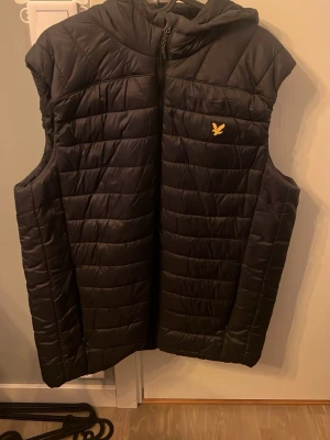 Svart dunväst från Lyle & Scott - Passar L Svart dunväst från Lyle & Scott med huva och gul broderad fågel på bröstet. Västen har quiltad design och dragkedja framtill. Perfekt för lager-på-lager och snygg till hoodie eller tröja. Klassisk och stilren look.