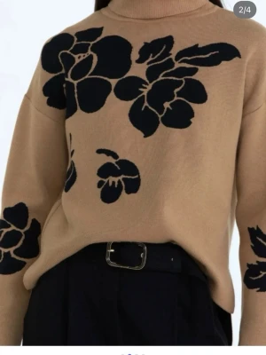 Beige polotröja med svarta blommor - Säljer en beige polotröja med stora svarta blommor som mönster. Tröjan har lång ärm och är stickad med ribbad polokrage och muddar. Perfekt för dig som gillar coola detaljer och vill ha något som sticker ut i garderoben.