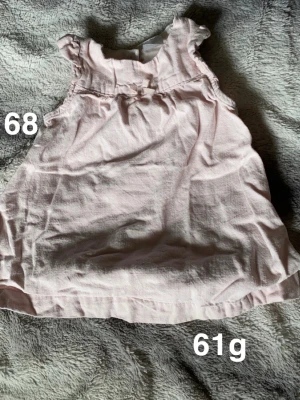 Ljusrosa ärmlös blus med rosett - Supersöt ljusrosa blus, ärmlös med små volangdetaljer vid axlarna och en liten rosett framtill. Perfekt för varma dagar och enkel att matcha med andra plagg.🌸 Fint använt skick. Storlek 68