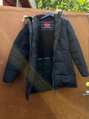 Svart lång pufferjacka Zavetti Canada - Säljer en svart lång pufferjacka från Zavetti Canada med huva och fuskpälsdetalj. Jackan har dragkedja framtill, stora fickor och quiltad insida. Perfekt för kalla dagar och riktigt snygg till vinterlooken.