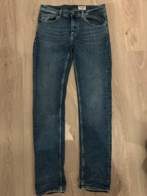 Blå jeans med raka ben från Tiger of Sweden - Snygga blå jeans från Tiger of Sweden med klassisk femficksdesign och raka ben. Jeansen har en mörk tvätt med subtila slitningar och kontrastsömmar. De är tillverkade i mjukt jeansmaterial och har en normal passform med medelhög midja. Perfekt för dig som gillar stilrena och tidlösa jeans.