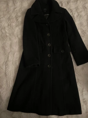 Svart lång kappa från Lindex - Säljer en svart kappa från Lindex med klassisk krage och fem stora knappar framtill. Jackan har lång ärm, markerad midja och två fickor med knappdetalj. Materialet är ullblandning som håller dig varm under kyliga dagar. Perfekt för dig som gillar stilrena och tidlösa plagg.
