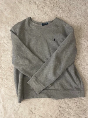 Grå sweatshirt från Polo Ralph Lauren - Säljer en stilren grå sweatshirt från Polo Ralph Lauren i storlek L 14-16. Tröjan har rund hals, lång ärm och klassisk broderad logga på bröstet. Mjuk bomullskvalitet som är skön att ha på sig. Perfekt för chill dagar eller när du vill ha en enkel look. Missa inte att det är barnstorlek!