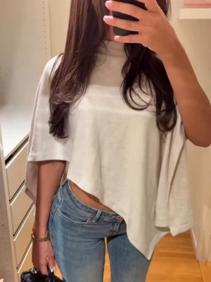 Vit croppad ponchotopp - Säljer en vit croppad topp med ponchoinspirerad passform och asymmetrisk nederkant. Toppen har hög hals och är tillverkad i ett mjukt, tunt material som faller snyggt. Perfekt för dig som gillar en avslappnad men trendig stil.