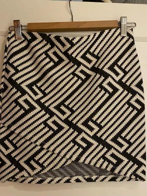 Svartvit rutig kortkjol - Säljer en svartvit kortkjol med ett coolt grafiskt rutmönster. Kjolen har en rak passform och är tillverkad i ett tjockare tyg som ger struktur. Perfekt för dig som gillar att sticka ut med mönster och vill ha en trendig look.