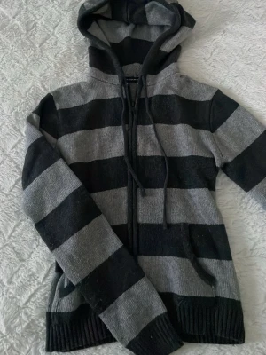 Zip från Brandy Melville - Säljer en stickad hoodie från Brandy Melville med breda svarta och grå ränder. Tröjan har huva, snörning och dragkedja framtill samt långa ärmar och ribbade muddar. One size men passar XS-M! 💘💘