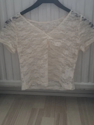 Vit spetsblus med v-ringning - Säljer en ljusvit blus i spets med korta ärmar och v-ringad hals. Blusen har en snygg rynkning framtill och är croppad i modellen. Spetsen har ett blommigt mönster som ger en romantisk vibe. Perfekt att styla med jeans eller kjol.