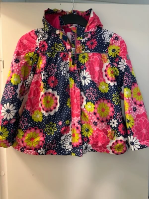 Blommig vårjacka  med rosa foder strl: 122 - Färgglad vindjacka från ME TOO med stort blommönster i rosa, vitt och limegrönt på mörkblå botten. Jackan har huva, dragkedja framtill, rosa innerfoder och snörning i midjan för bästa passform. Perfekt för vår och höst när du vill sticka ut.