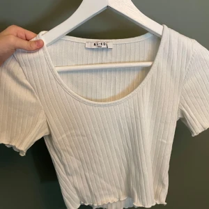 Vit ribbad topp  - Superfin vit topp från NA-KD i ribbad bomullsmix. T-shirten har korta ärmar, rund halsringning och vågig kant nertill. Perfekt till jeans eller kjol för en clean och fräsch look.