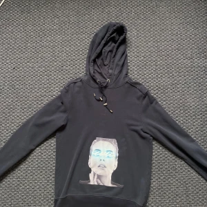 Limitato hoodie - Blå hoodie från Limitato med ett stort grafiskt tryck av ett ansikte på framsidan där ögonen är täckta av blå neonliknande streck. Tröjan har huva med snörning och långa ärmar. 