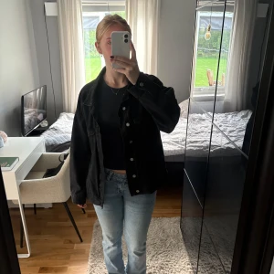 Svart jeansjacka med knappar - Säljer en svart jeansjacka med klassisk krage och silverfärgade knappar framtill. Jackan har två bröstfickor med knapp och en loose passform som ger en chill vibe. Perfekt att slänga över en t-shirt eller hoodie för en cool look.