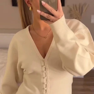 Krämvit kofta med v-ringning och knappar - Superfin krämvit kofta med djup v-ringning och snygga klädda knappar framtill. Modellen är figurnära med långa ärmar och lätt oversized axlar. Perfekt att styla med jeans eller kostymbyxor för en clean look.