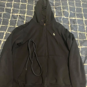 Svart hoodie från Polo Ralph Lauren - Svart hoodie från Polo Ralph Lauren med dragkedja, huva och snörning. Klassisk broderad logga på bröstet och stora fickor framtill. Tillverkad i mjuk bomull, perfekt för chill dagar eller när du vill ha en clean streetstyle.