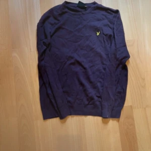Mörklila tröja från Lyle & Scott - Snygg mörklila långärmad tröja från Lyle & Scott med deras klassiska gula fågel-logga på bröstet. Tröjan har rund hals och är gjord i mjuk bomull, perfekt för chill dagar. Passar dig som gillar stilrena och enkla plagg med en liten detalj.