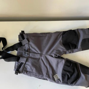 Grå termobyxor från On The Peak - Säljer ett par grå täckbyxor från On The Peak med svarta förstärkningar på knän och bak. Byxorna har justerbara hängslen och praktiska kardborreband i midjan. Perfekta för kalla dagar och vinteraktiviteter. Snygg logga på benet.