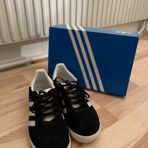 Adidas Gazelle J svart/vit strl 38 - Säljer ett par klassiska Adidas Gazelle J sneakers i svart mocka med vita detaljer och vita ränder på sidorna. Skorna har platt sula, rund tå och snörning. Ikonisk Adidas-logga på hälen och vit yttersula. Perfekta för dig som gillar retrostil och streetwear. Nästan helt nya använt 2 gånger säljer för dom inte kommer till användning, Du får med låda storlek 38💞 Skriv om ni har frågor eller bilder