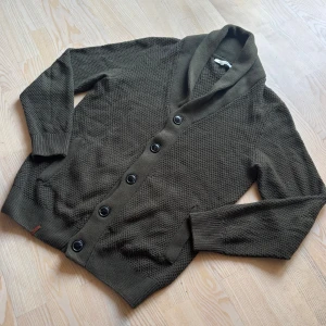 Stickad Cardigan - Säljer en mörkgrön stickad kofta med sjalkrage och stora svarta knappar. Koftan har två fickor framtill och är långärmad med ribbade muddar. Perfekt för lager-på-lager och chill höststil.