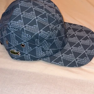 Blå mönstrad keps från Lacoste - Snygg blå keps från Lacoste med geometriskt mönster i ljusblått. Klassisk fempanelsdesign med justerbar rem bak och metallöljetter på sidorna. Ikoniska Lacoste-krokodilen broderad på sidan. 