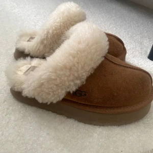 Bruna UGG tofflor med fårull - Mysiga bruna UGG tofflor med fluffig vit fårullskant och insida. Ovandel i mocka. Perfekt nu till hösten/vintern 🫶🏻står storlek 36 på lappen men passar perfekt på mig som vanligtvis har 37/38❣️ fint skick men såklart finns det lite tecken på användning men inte mycket , det som är sysn på bilderna ☺️ hör gärna av er vid frågor !