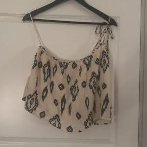  mönstrad linnetopp med knyt - Säljer en beige linnetopp med svart grafiskt mönster. Toppen har tunna axelband och en snygg knytning på ena axeln. Använd några gånger men väldigt bra skick.