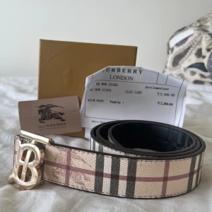 Burbery bälte - Snyggt Burbery bälte med klassiskt rutigt mönster i beige, svart och röd. Bältet har ett stort, guldigt TB-spänne framtill och är gjort i skinn. Perfekt för att lyfta vilken outfit som helst med en lyxig touch och ikonisk design.