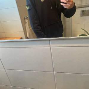 Stone Island hoodie - Svart hoodie från Stone Island med klassisk patch på vänster ärm och logga på bröstet. Tröjan har huva med snörning och en stor magficka. Perfekt för dig som gillar streetwear och vill ha en clean look.