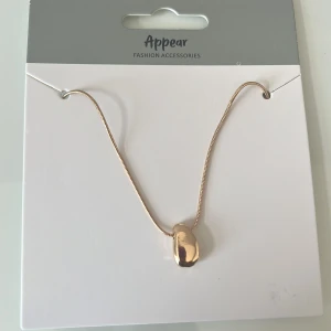 Guldigt halsband från Appear - Snyggt halsband i guldton med en slät, organisk formad berlock. Kedjan är tunn och smidig med justerbar längd. Perfekt för dig som gillar minimalistisk och modern stil. Passar till alla dina favoritoutfits och ger en lyxig touch. Aldrig använd